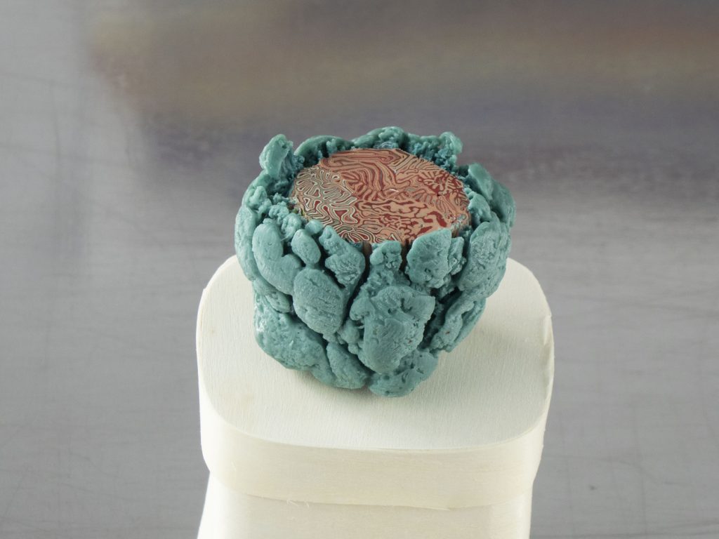 Small encaustic sculpture of a teal lumpy sphere cradling a circular disc patterned with intricate red labyrinthine lines.