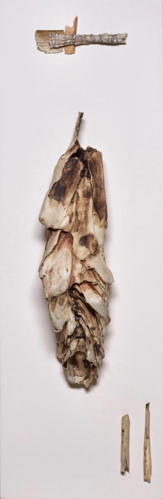 Ceramic and wood assemblage of layered husk-like forms hanging vertically on a white background.
