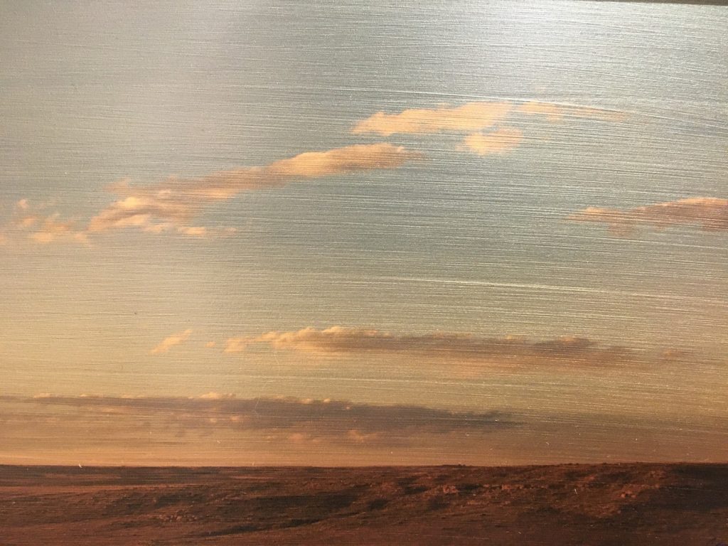 Photograph on painted steel of a flat open landscape under a wide evening sky with wispy warm-toned clouds.