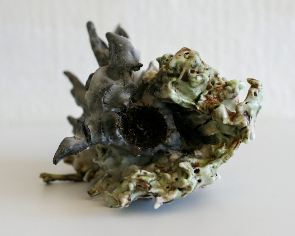 Small ceramic sculpture with jagged spikes and pitted surfaces, glazed in gray, celadon green, and rust brown.