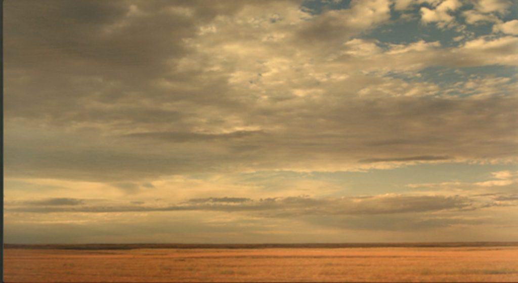 Photograph of a flat golden prairie under a vast cloud-filled sky with patches of pale blue.