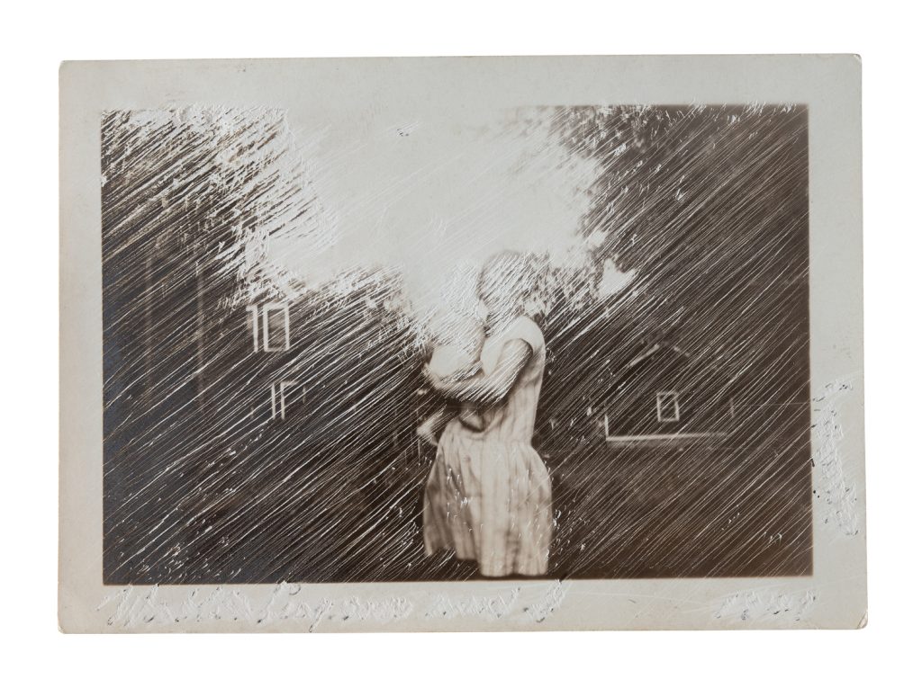 Found photograph of a standing figure in a dress, heavily scratched with diagonal incisions that obscure most of the image.