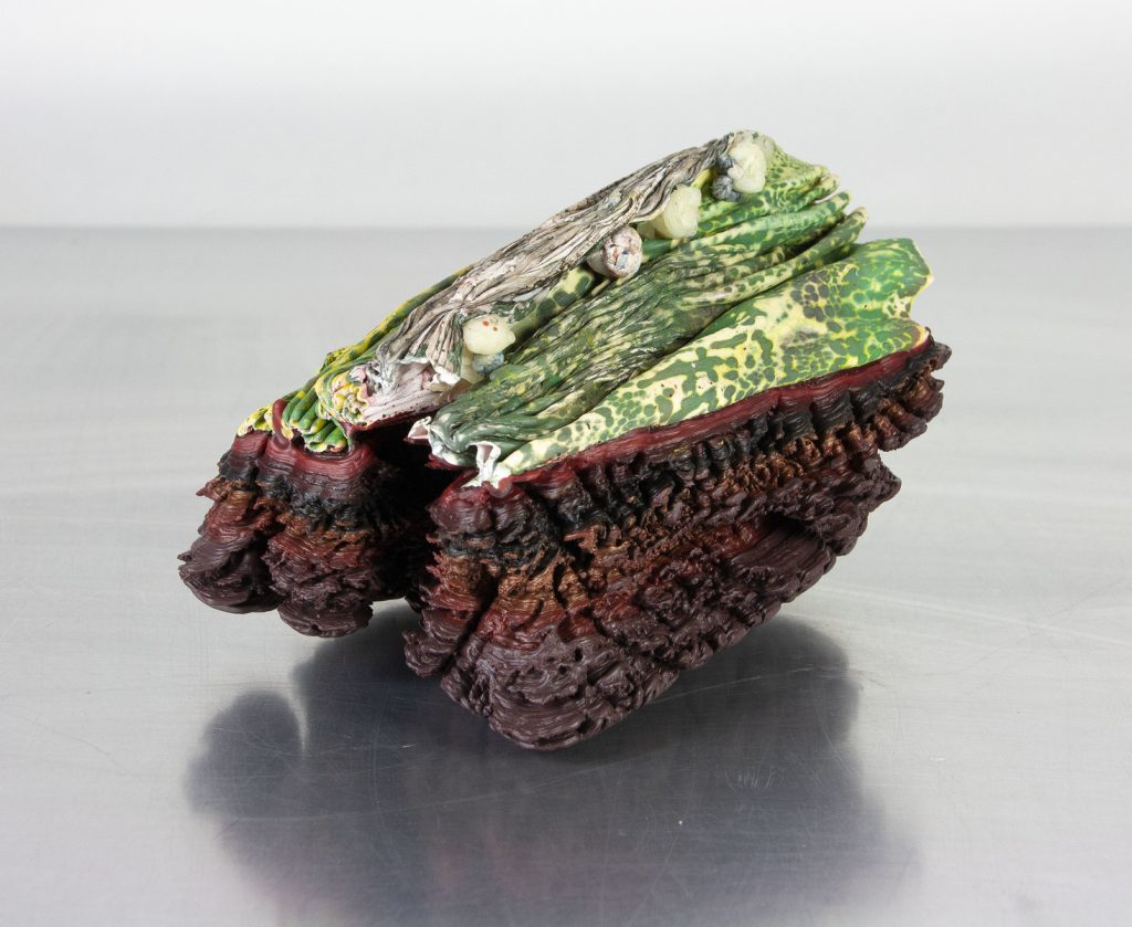Pigmented beeswax sculpture resembling a gnarled organic form with dark reddish base and layered green leaflike upper surfaces.
