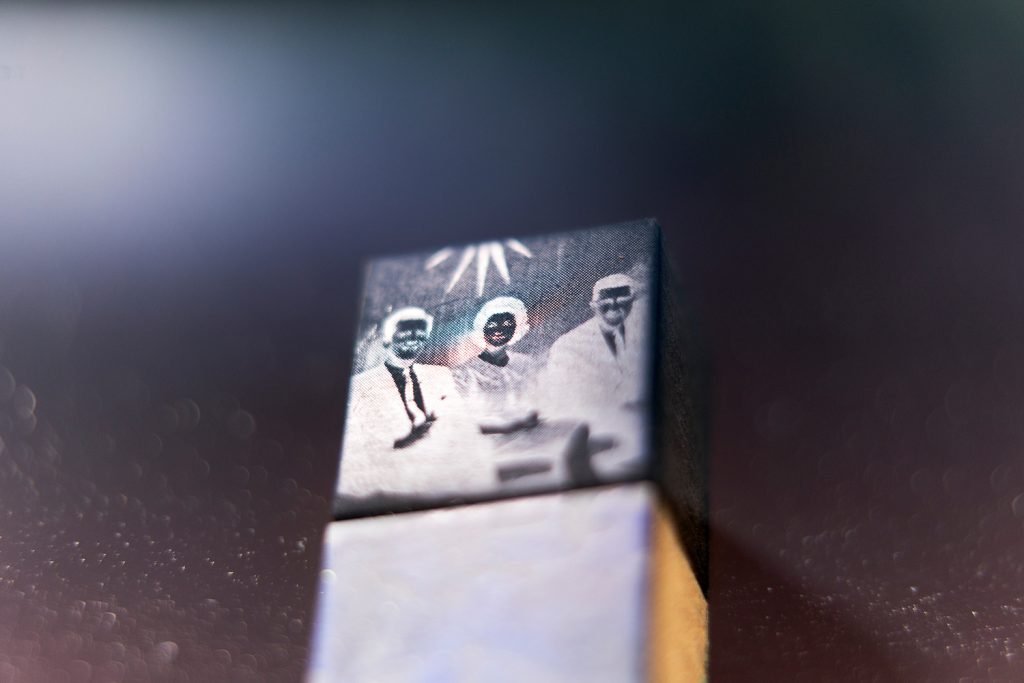 Pigment print of a small block displaying a black-and-white image of three figures with obscured faces and radiating light rays.