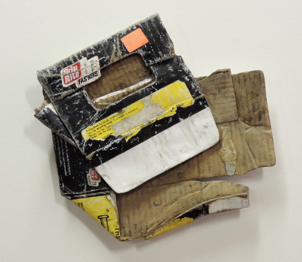 Wall-mounted acrylic on wood sculpture of a crushed Grip Rite Fasteners cardboard box with yellow and black labeling.