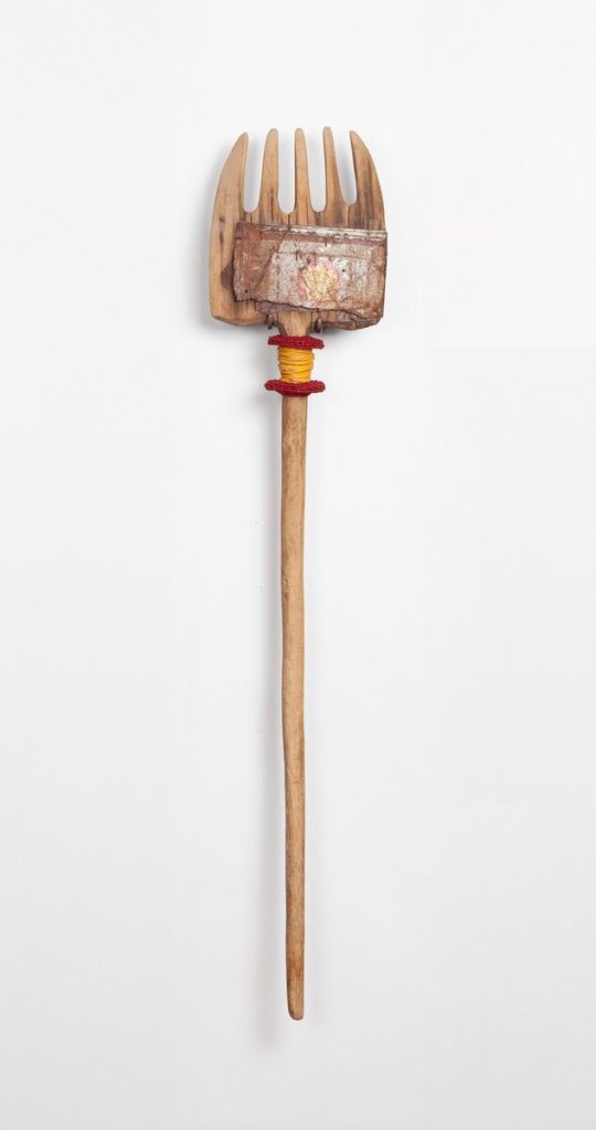 A weathered wooden hay fork with red and yellow waxed linen binding mounted vertically against a white wall.
